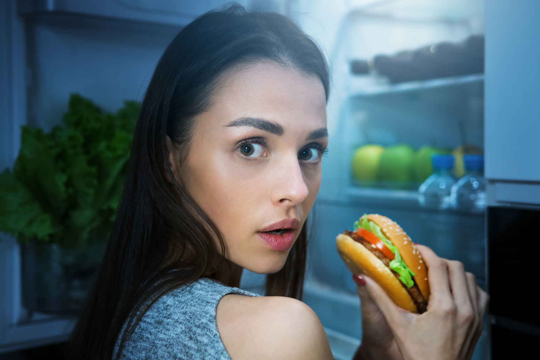 Hungry woman eating burger at night near fridge